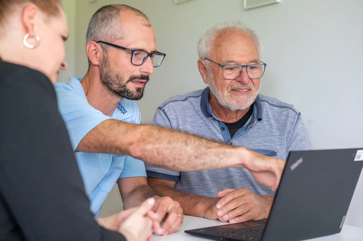 Rehakoordinator im Gespräch mit Patient und Angehöriger, erklärt etwas am Laptop.