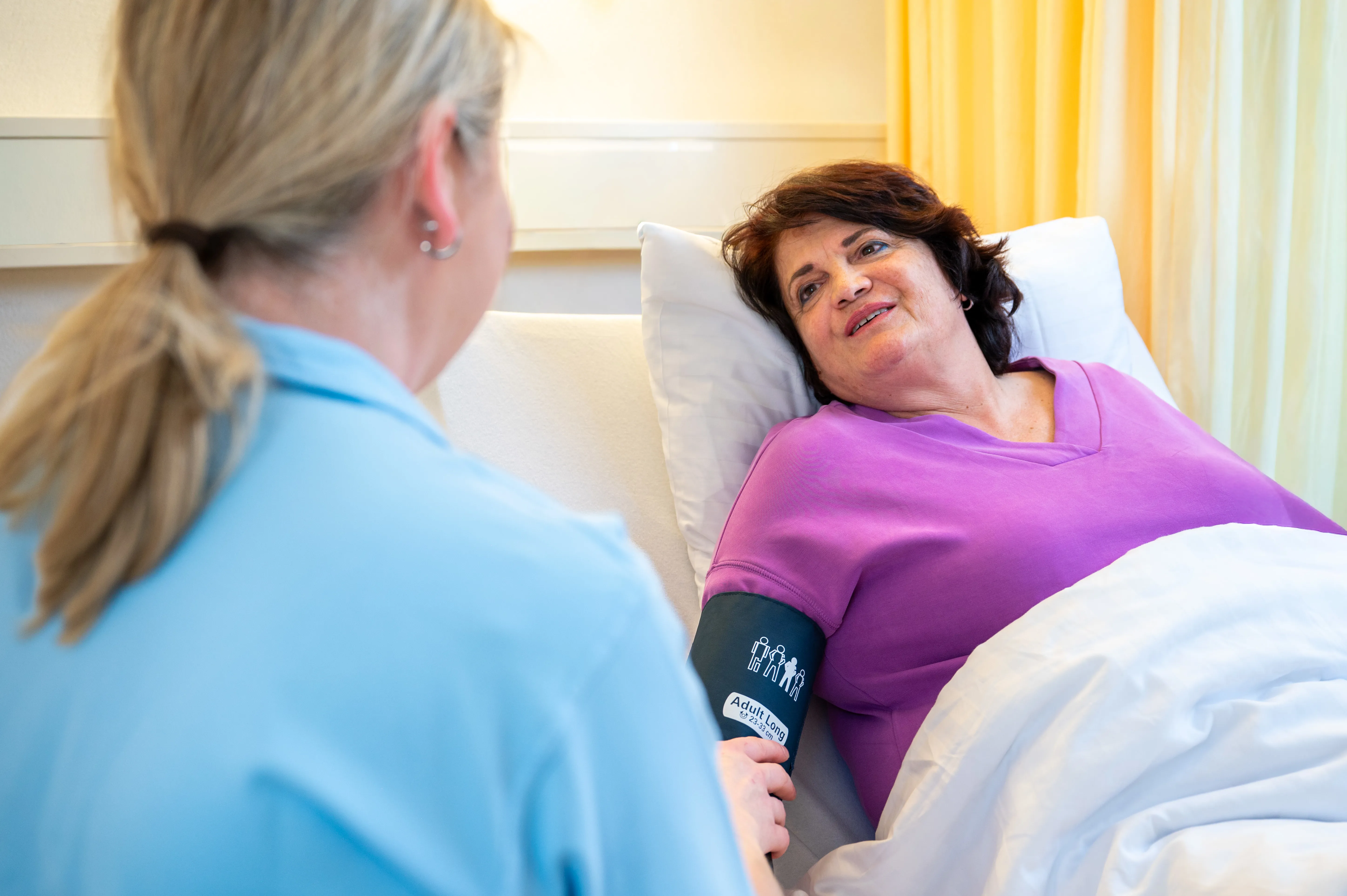 Eine Patientin liegt im Bett während eine Pflegefachfrau ihren Blutdruckt misst.