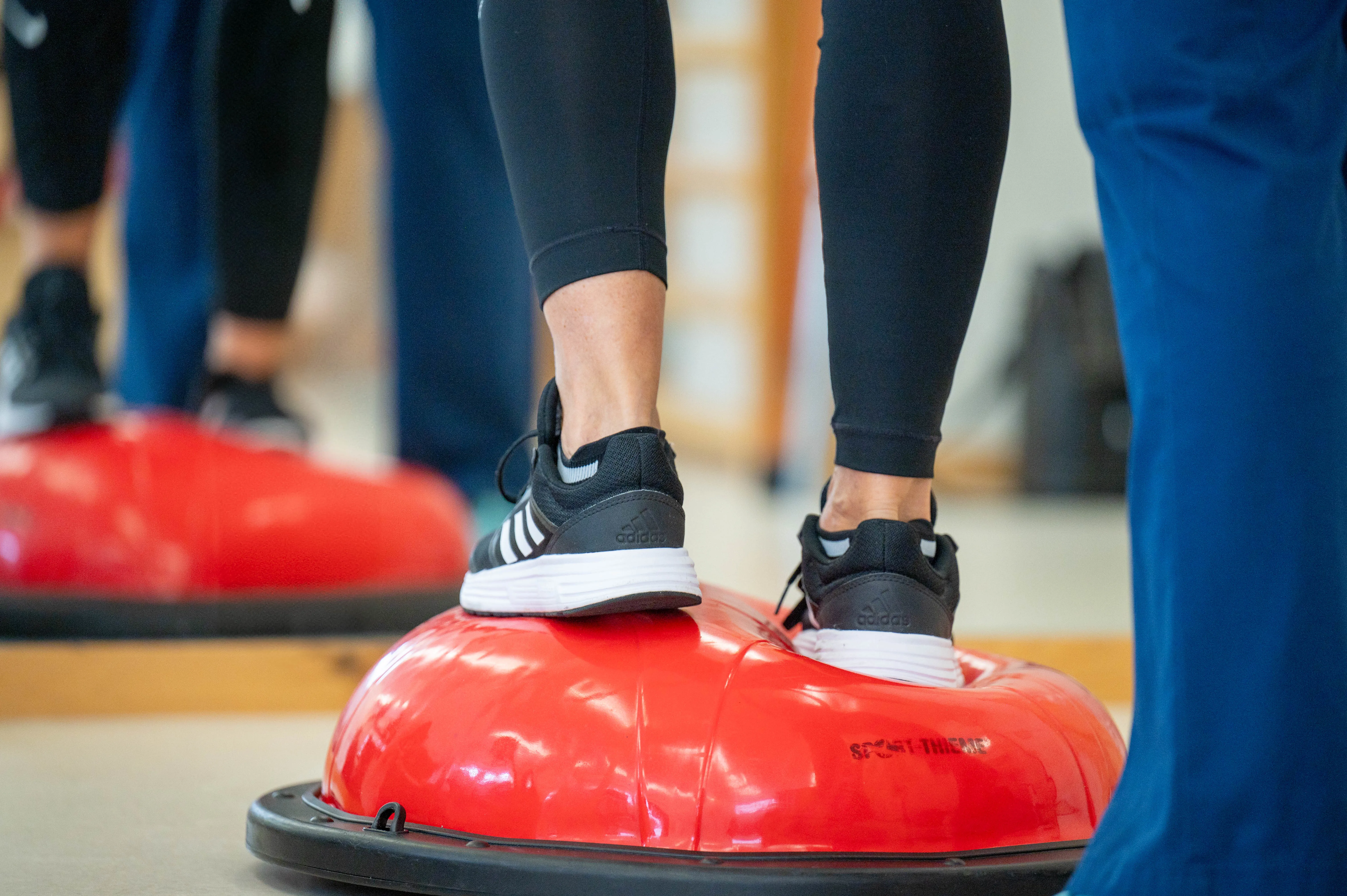 Beine einer Patientin während einer Übung auf einem halbrunden Ball, begleitet von einer Therapeutin.