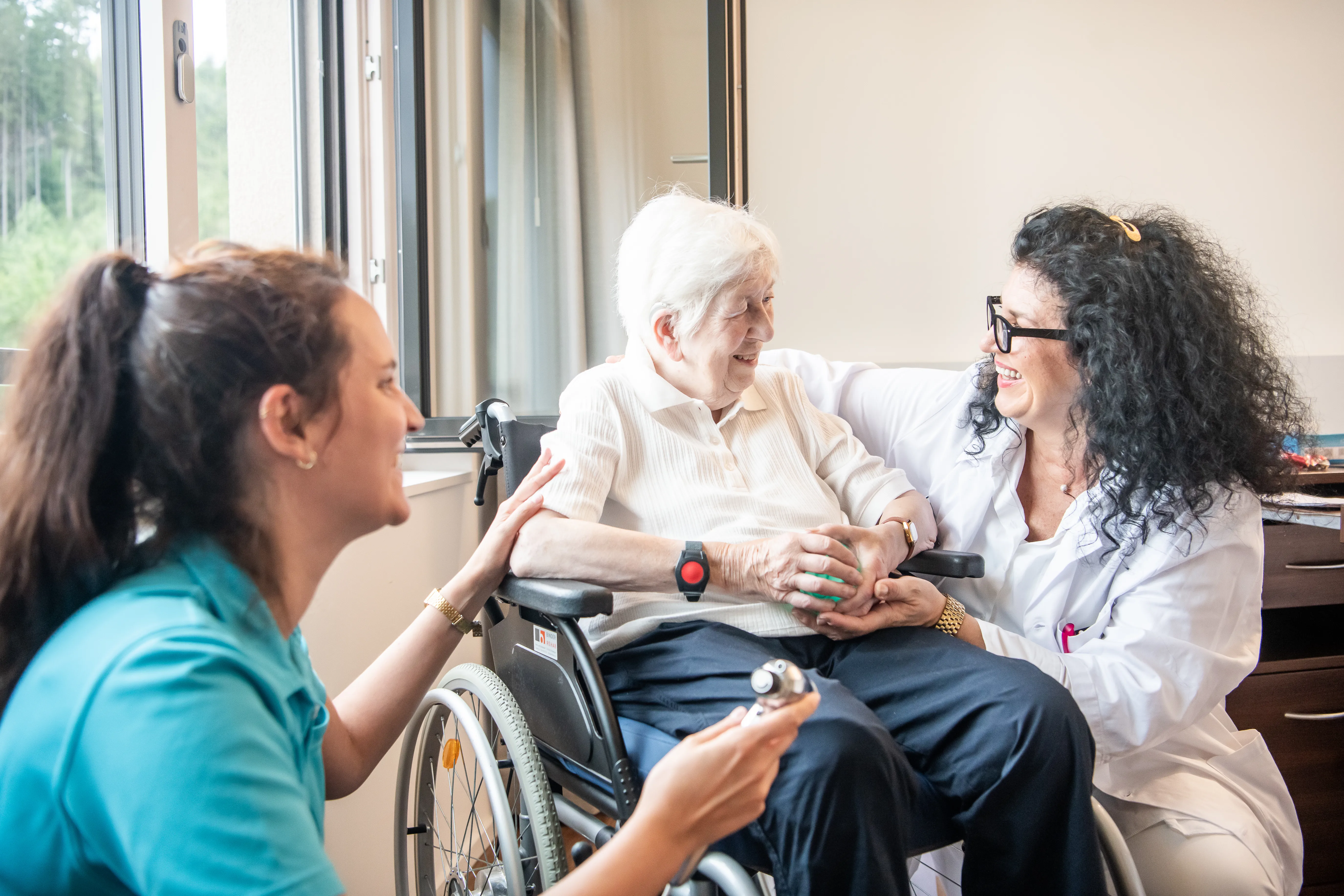 Eine Ärztin und eine Pflegefachfrau umsorgen eine ältere Patientin im Rollstuhl und lachen mit ihr.