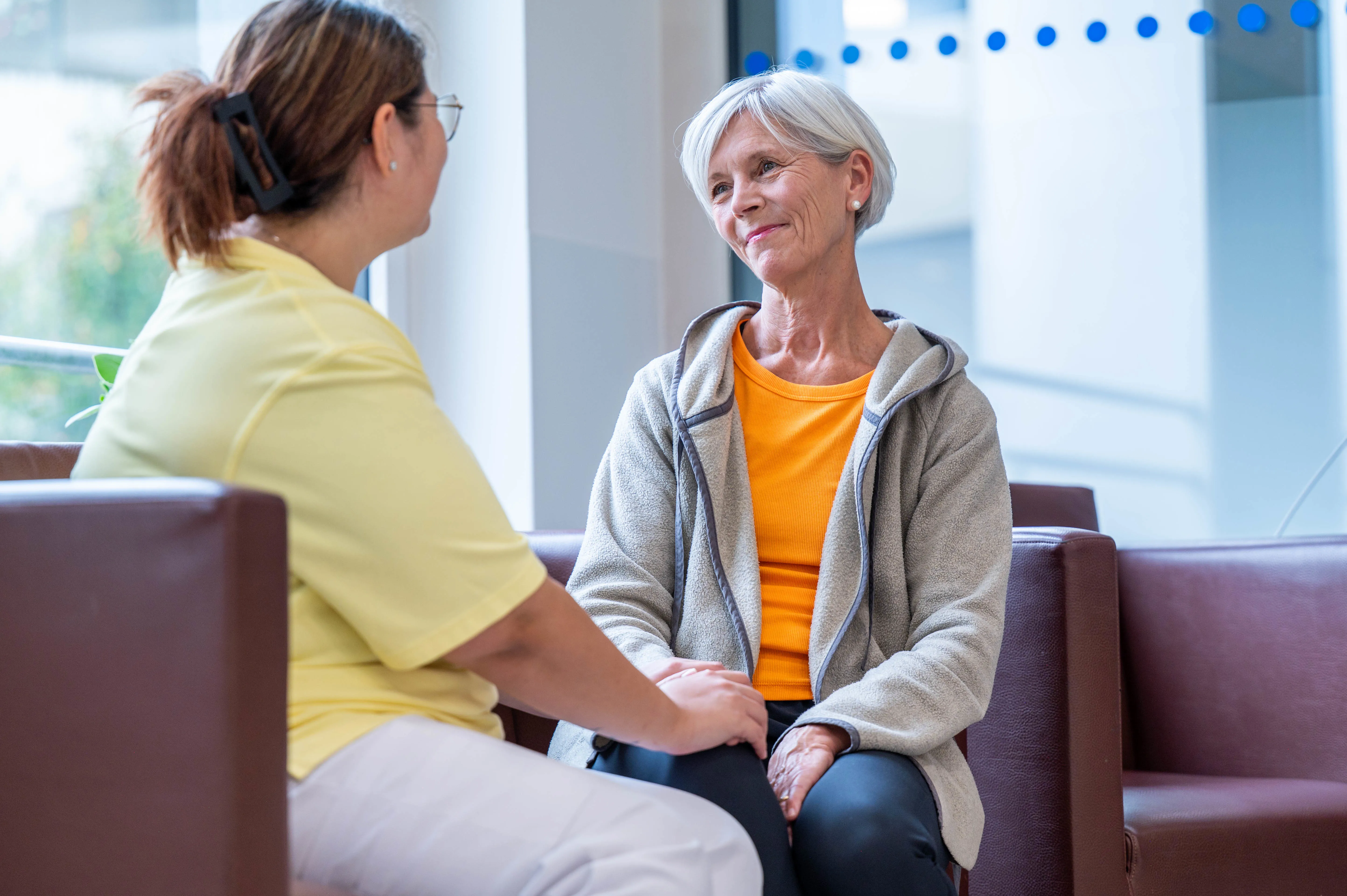 Ältere Dame im Gespräch mit Pflegefachfrau, die ihre Hand auf ihr Knie legt.