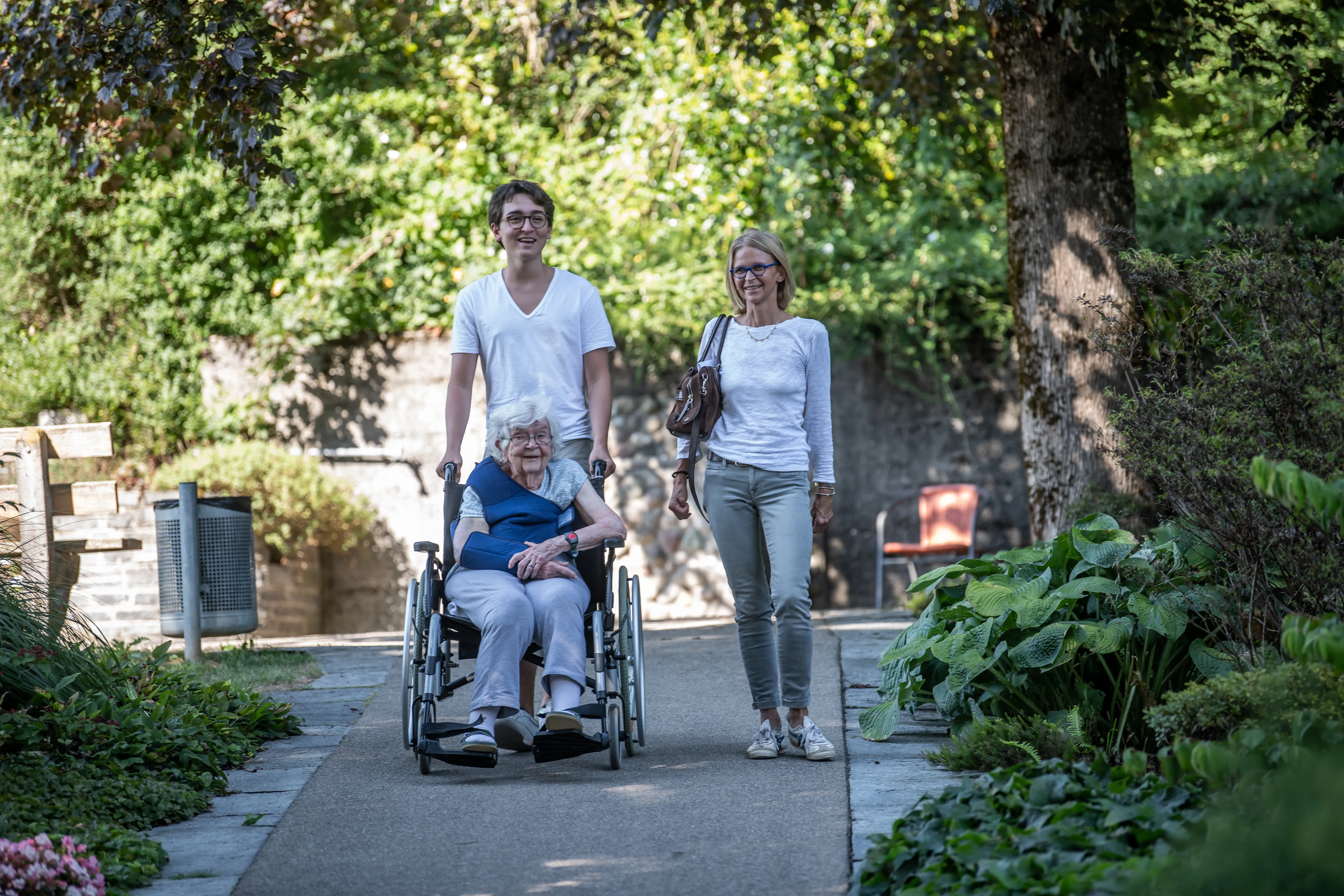 Ältere Patientin im Rollstuhl, begleitet von Enkel und Tocher bei einem Spaziergang im Park.